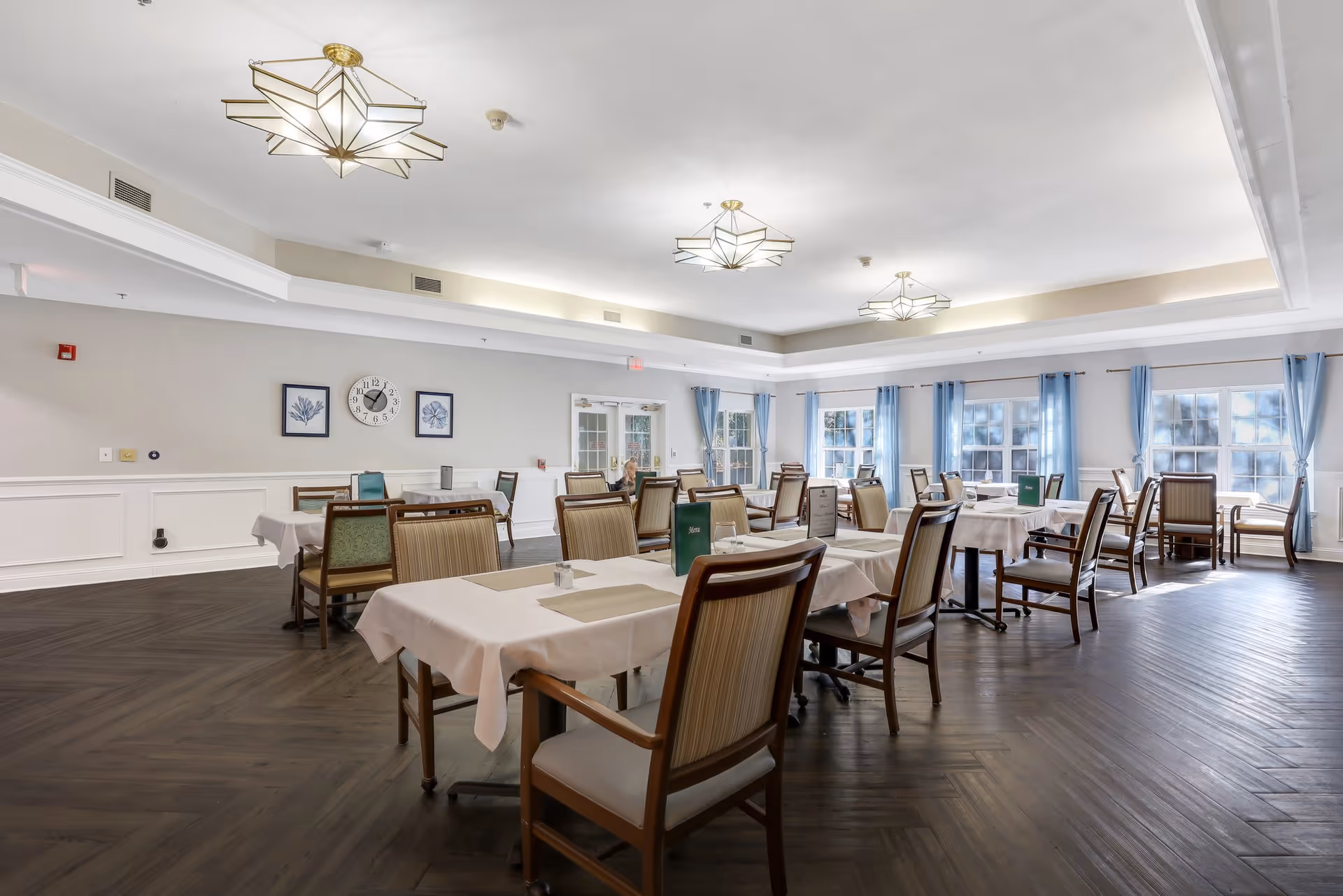 A spacious dining room with multiple tables covered in white tablecloths and set with placemats and menus. The room features large windows with blue curtains allowing natural light to fill the space, elegant ceiling lights, and a wall clock with framed artwork on the wall.