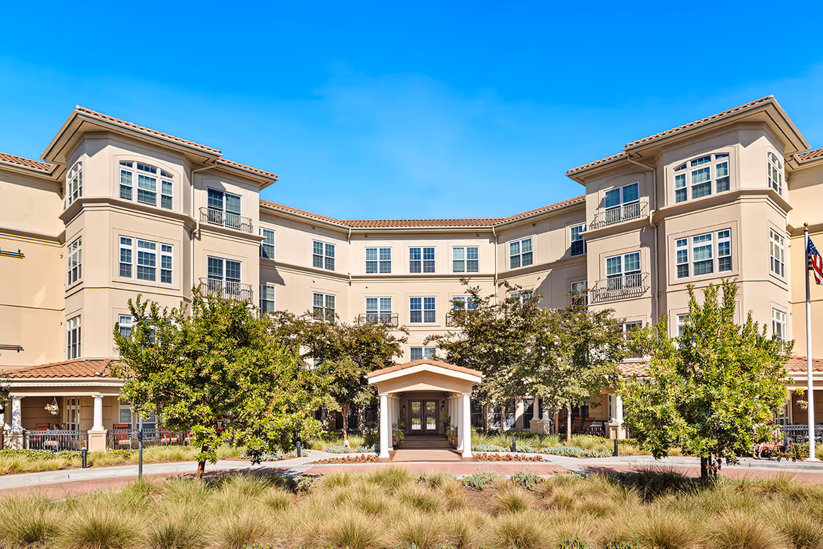 Front exterior view of a multi-story senior living facility building with beige walls, multiple windows, and a tiled roof under a clear blue sky. The entrance has a covered walkway supported by white columns, surrounded by landscaped greenery and trees.