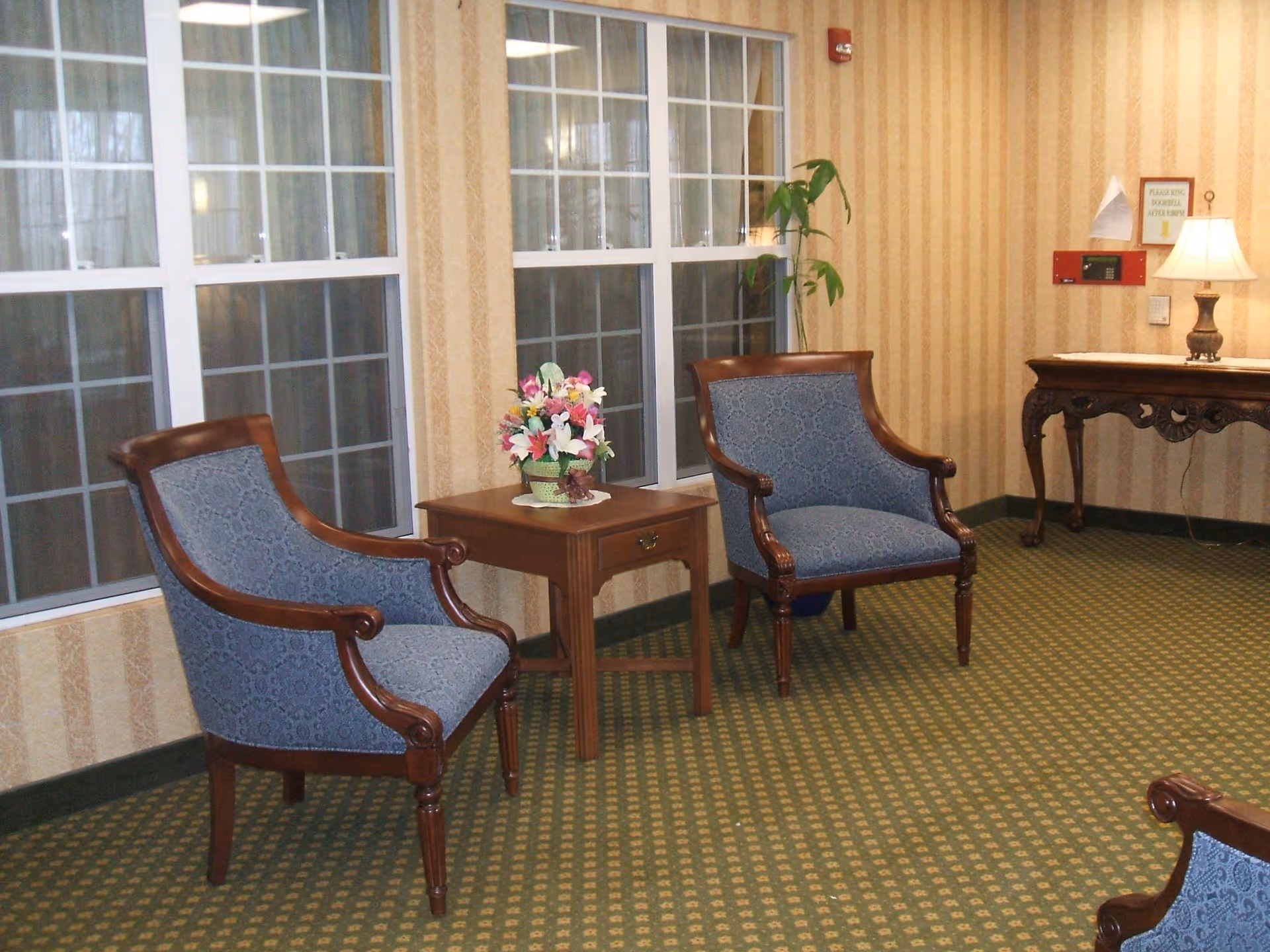 A sitting area with two blue upholstered wooden armchairs placed on either side of a wooden side table with a floral arrangement. The room has large windows with white frames, striped beige wallpaper, and a green patterned carpet. A wooden console table with a lamp and a small sign is visible in the background.