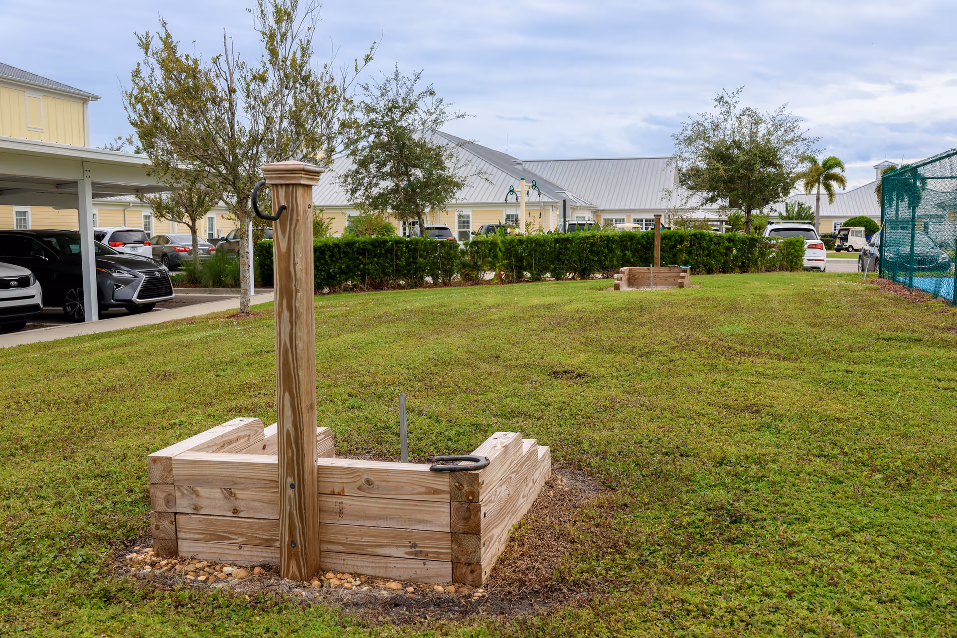 Outdoor grassy area with two wooden horseshoe game pits, surrounded by parked cars, trees, and buildings with metal roofs under a cloudy sky.
