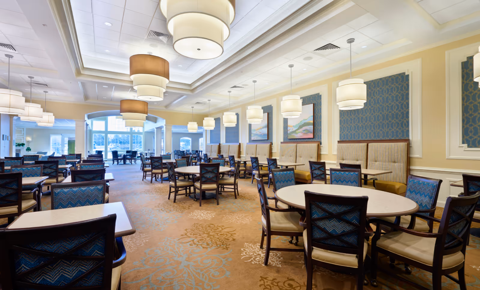 A spacious and well-lit dining room in a senior living community featuring multiple tables and chairs arranged neatly. The room has large windows allowing natural light, elegant hanging light fixtures, cushioned seating along the walls, and decorative wall panels with artwork.