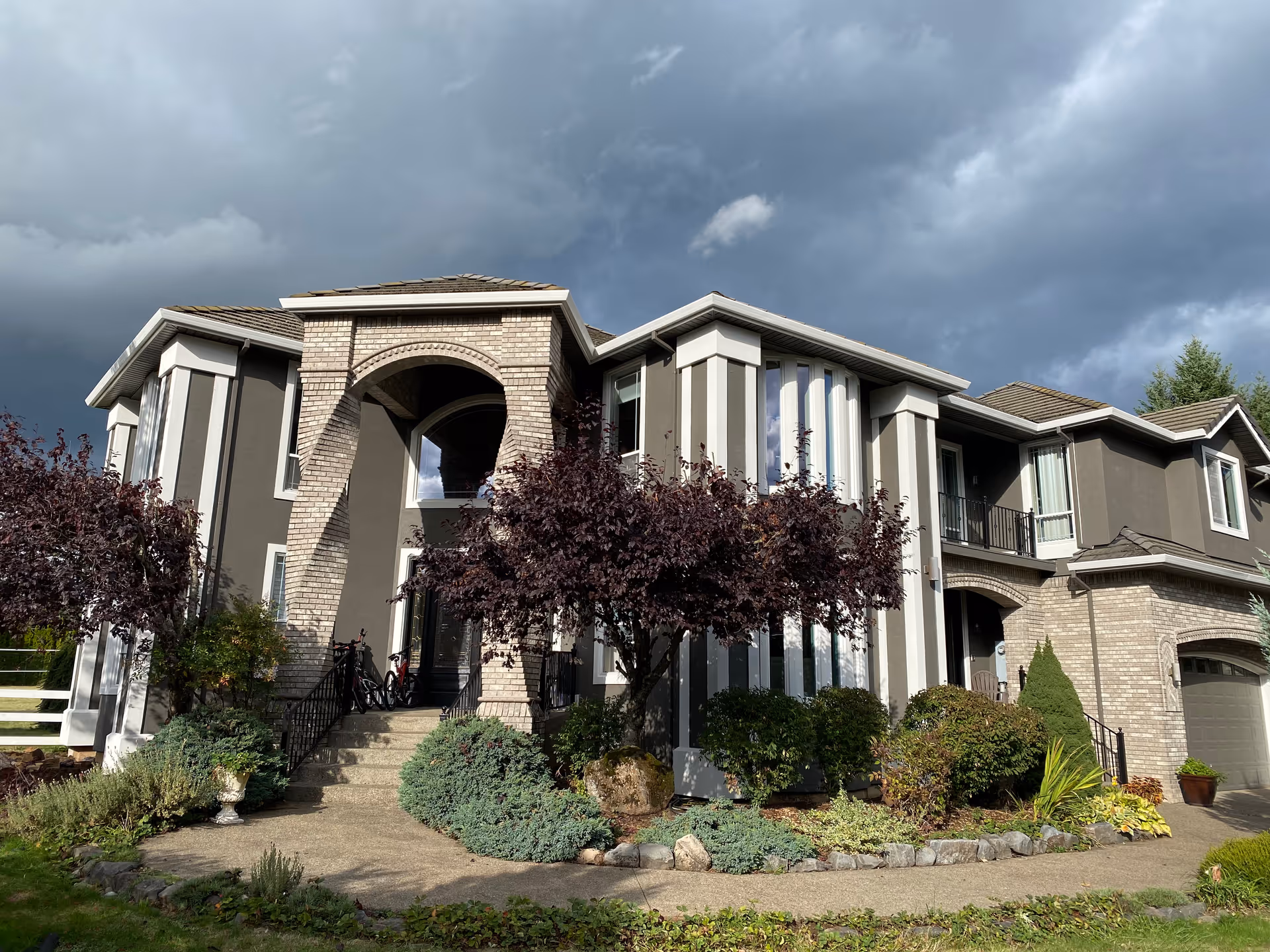 Two-story residential building with a brick arched entrance, landscaped front yard, and a dramatic cloudy sky overhead.