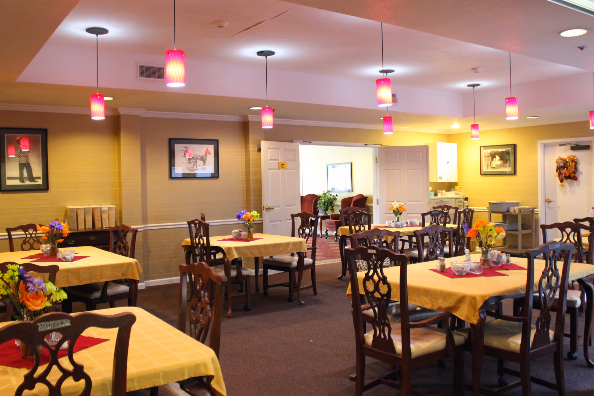 A dining room in a senior living community with multiple tables covered in yellow tablecloths and red placemats. Each table has a floral centerpiece. The room has wooden chairs, framed pictures on the walls, and several hanging pendant lights with red shades. An open doorway leads to a lounge area with armchairs.