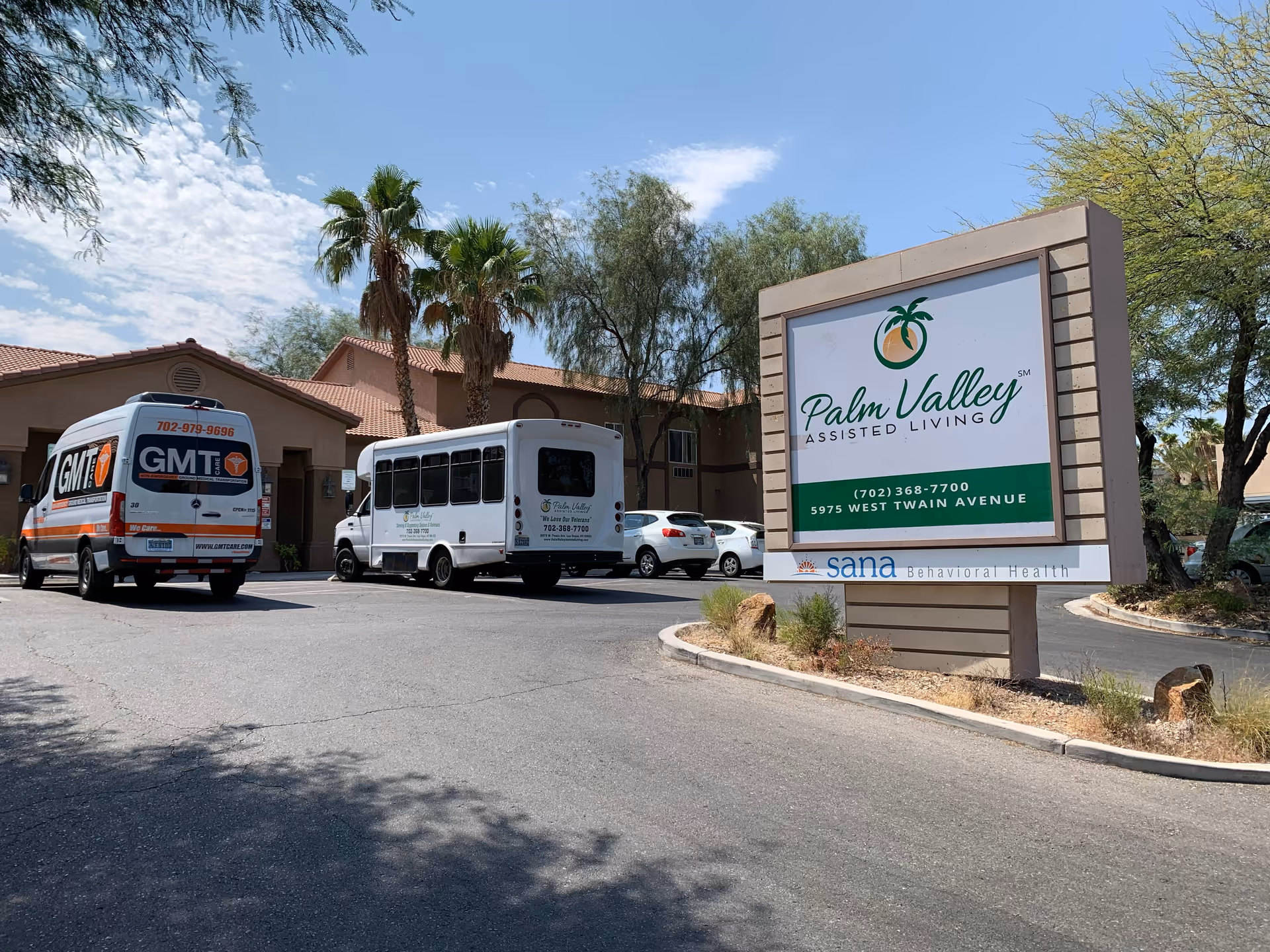 Exterior view of Palm Valley Assisted Living facility with a large sign displaying the facility name, phone number, and address. Two vehicles, including a shuttle bus and a van, are parked near the building entrance. Palm trees and desert landscaping are visible under a partly cloudy sky.