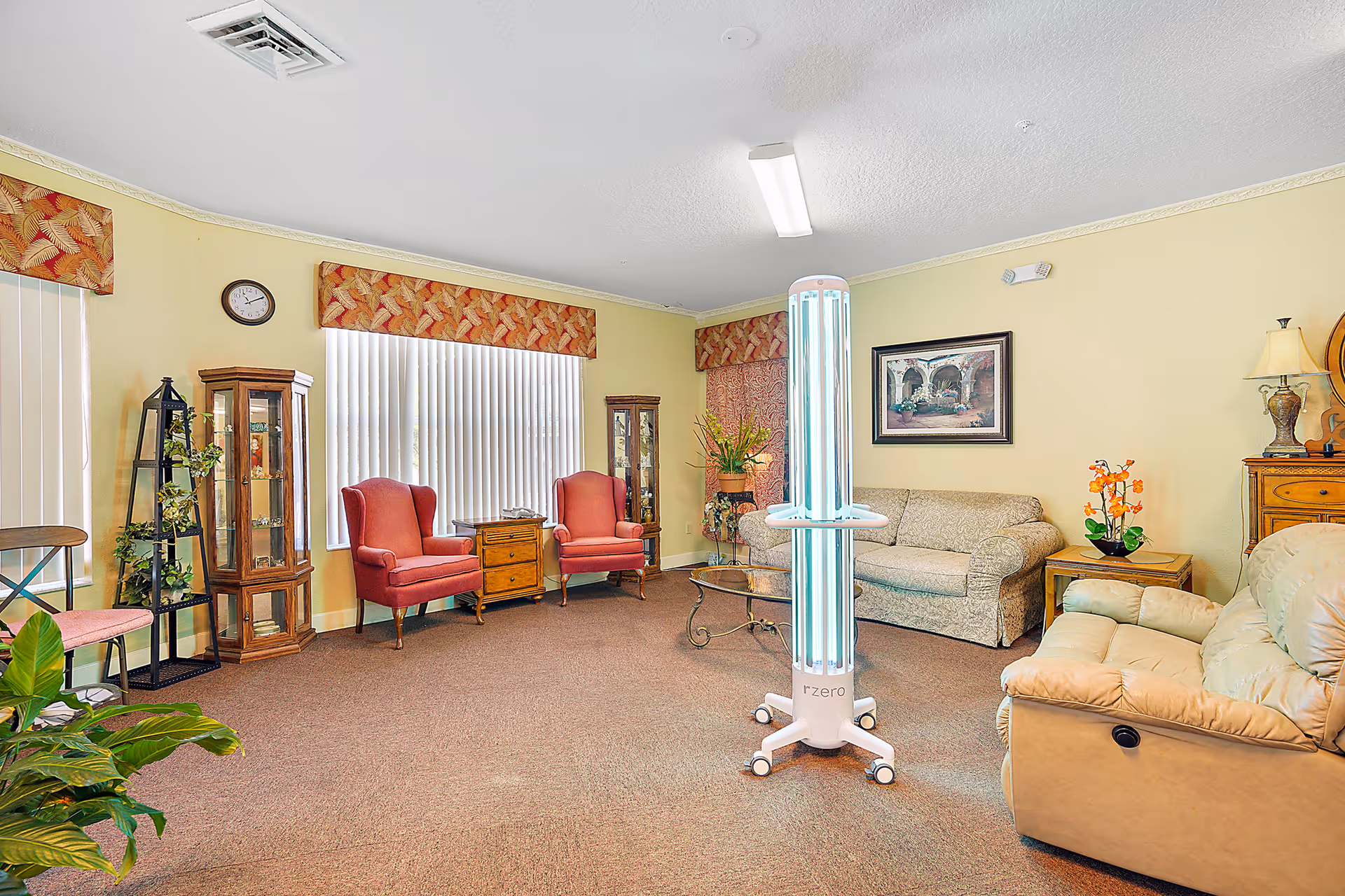 A cozy living room with beige walls and carpeted floor featuring two red armchairs, a beige sofa, a beige recliner, glass display cabinets, a small wooden side table, and a coffee table. There is a tall UV light sanitizer device in the center of the room. The windows have vertical blinds and patterned valances. Various plants and framed artwork decorate the space.