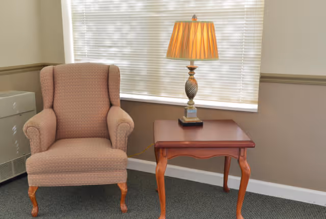 A wingback armchair beside a wooden side table holding a lamp in front of a window with closed blinds.