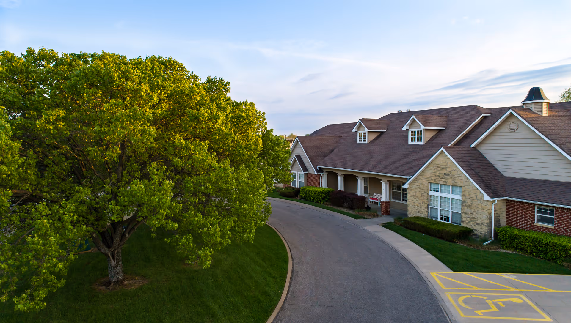A curved driveway leading to the front entrance of a single-story assisted living building with a large green tree on the lawn.
