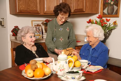 Three elderly women sitting and standing around a dining table in a cozy room. The table is set with a teapot, cups, a bowl of fruit, and a plate of cookies. One woman is standing and offering cookies to the two seated women, who are smiling and engaging with each other. The background shows wooden cabinets, a framed picture, and a vase with red flowers.