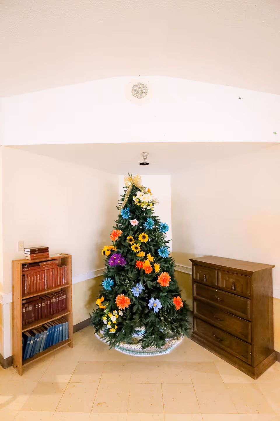 A corner of a room featuring a Christmas tree decorated with colorful artificial flowers instead of traditional ornaments. To the left of the tree is a small wooden bookshelf filled with books, and to the right is a wooden chest of drawers. The walls are light-colored and the floor is tiled.