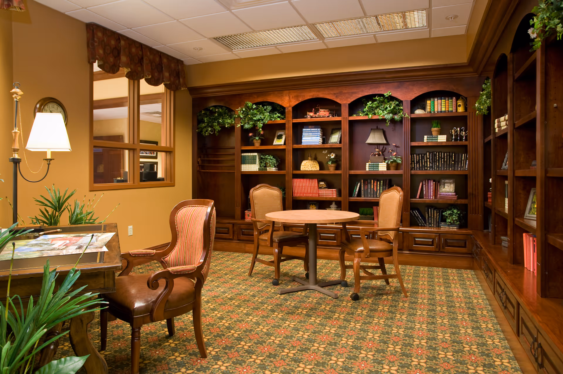 A cozy library or reading room with wooden bookshelves filled with books and decorative plants. There is a round wooden table with three upholstered chairs around it. A wooden desk with a lamp and some papers is visible on the left side. The room has a patterned carpet and warm lighting.
