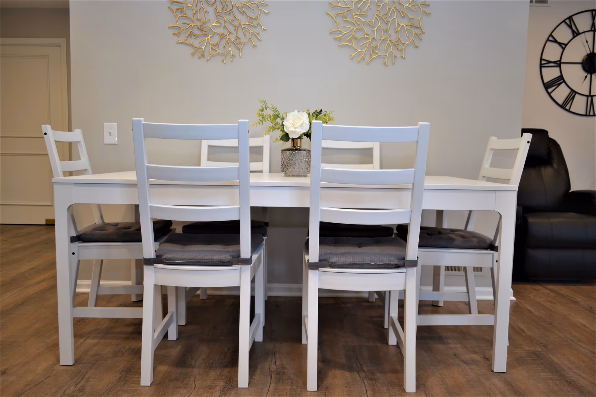 A white dining table with six white chairs, each with a gray cushion, placed on a wooden floor. A small floral arrangement in a vase is centered on the table. The background features a light gray wall with two decorative gold wall hangings and a large black clock on the right side. A black recliner chair is partially visible on the right.
