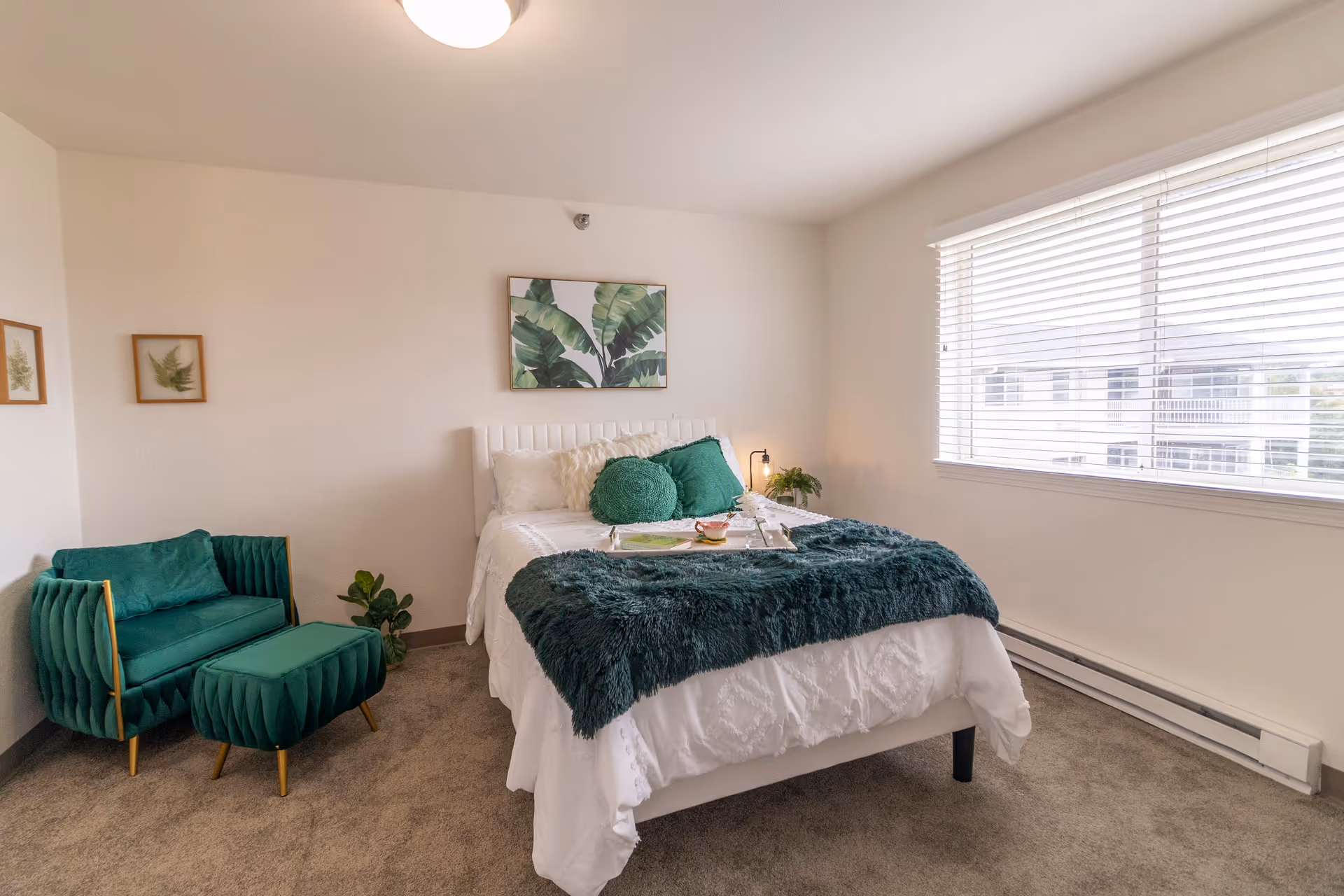 A bright bedroom with a white bed featuring green pillows and a dark green throw blanket. There is a matching green armchair with an ottoman to the left, a small plant on the floor, and framed botanical prints on the walls. A large window with blinds allows natural light to fill the room.