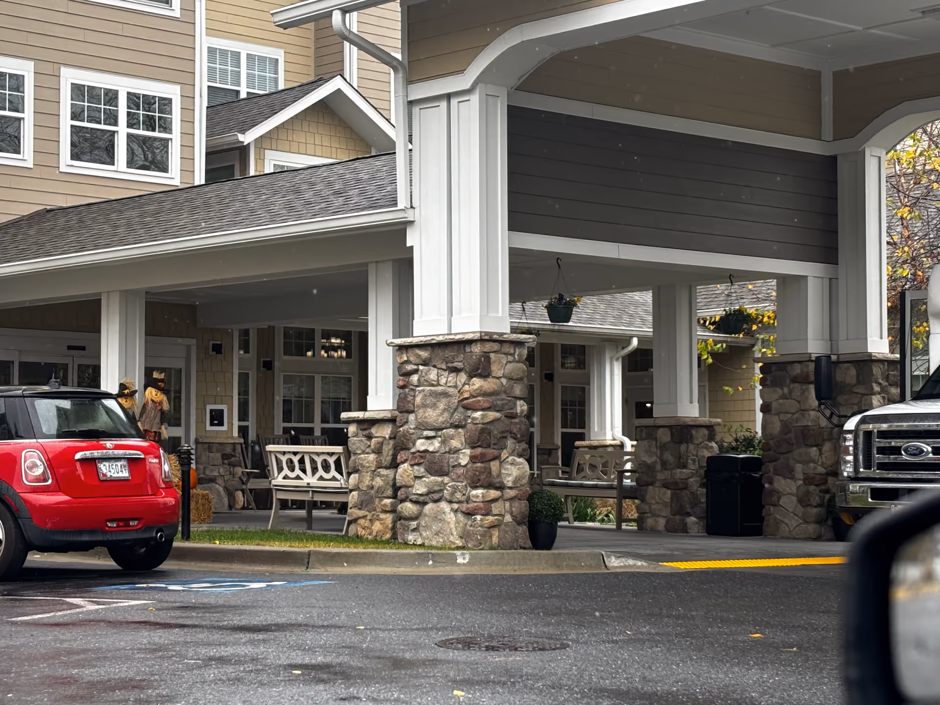 Entrance area of Wilshire Estates Gracious Retirement Living showing a covered drop-off zone with stone pillars and beige siding. There are benches and hanging plants under the covered area, a red Mini Cooper parked on the left, and a silver truck partially visible on the right.