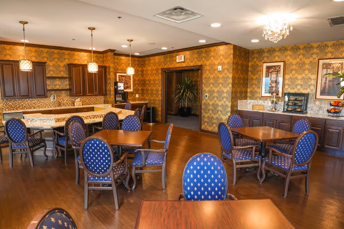 A dining room in a senior living facility with several wooden tables and blue upholstered chairs arranged neatly. The room features warm yellow patterned wallpaper, dark wood cabinetry, and a countertop area with pendant lights hanging above. There are framed pictures on the walls and a chandelier providing additional lighting.