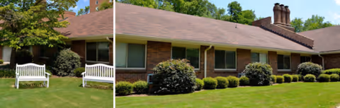 Exterior view of a single-story brick building with a brown shingled roof, surrounded by green grass, bushes, and trees. Two white benches are placed on the lawn in front of the building.