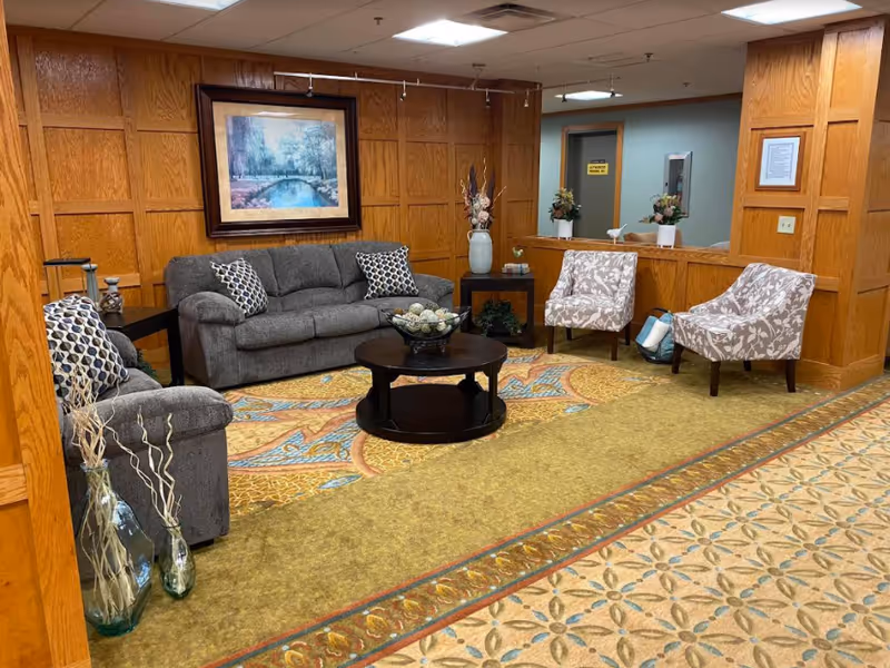 A cozy seating area in a senior living facility with a gray sofa, two patterned armchairs, a round wooden coffee table with decorative items, and wooden paneled walls. There is a large framed painting above the sofa and decorative vases with dried plants on the floor and side tables. The carpet has a colorful, intricate pattern.