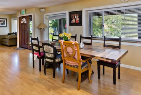 A bright dining area with a wooden table surrounded by six mismatched chairs, some with cushions. There are large windows with blinds letting in natural light and a door leading outside. The room has light-colored walls and wooden flooring, with a small living area visible in the background.
