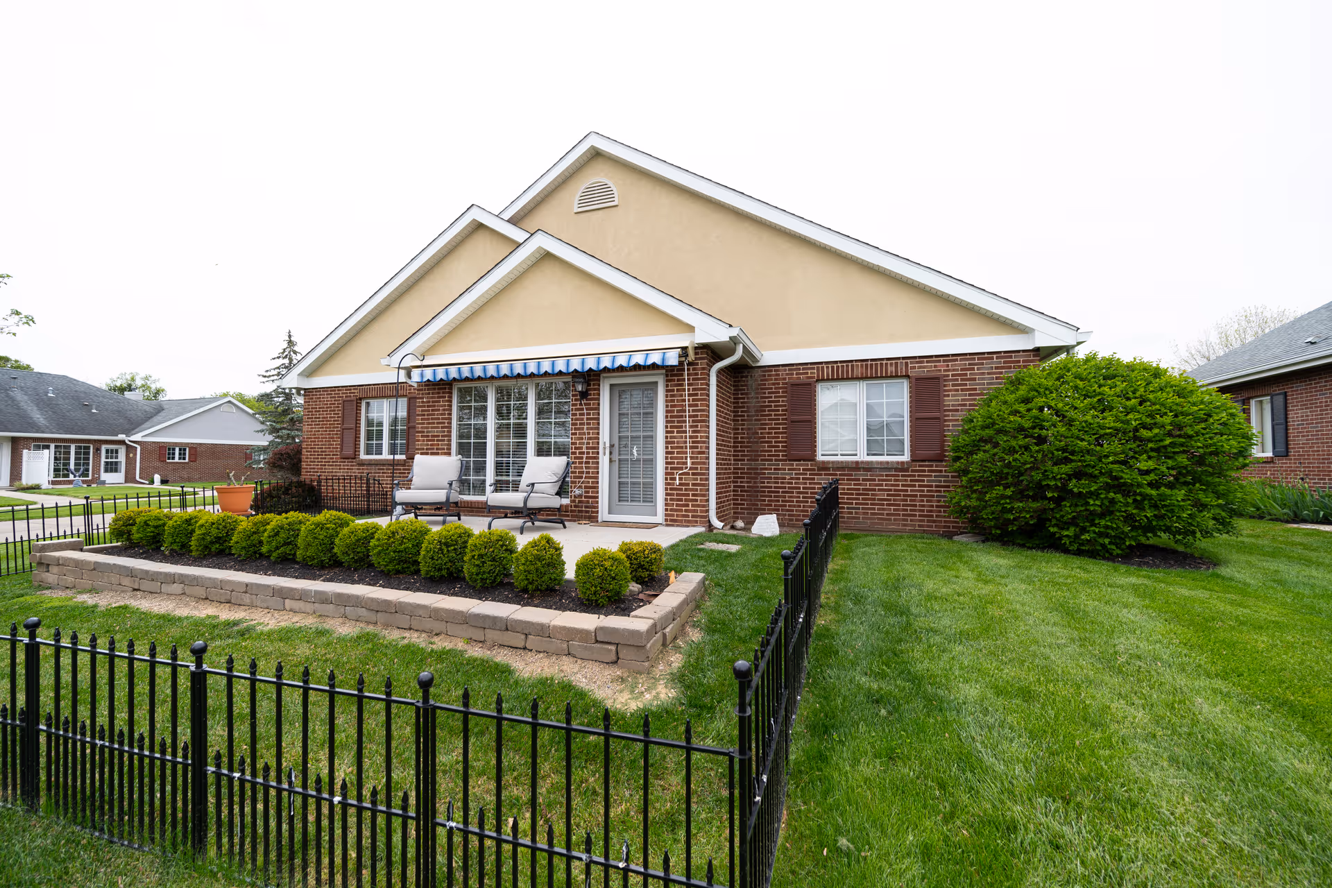 Single-story brick and stucco senior living cottage with a small fenced front patio, patio chairs under a striped awning, and manicured shrubs and lawn.