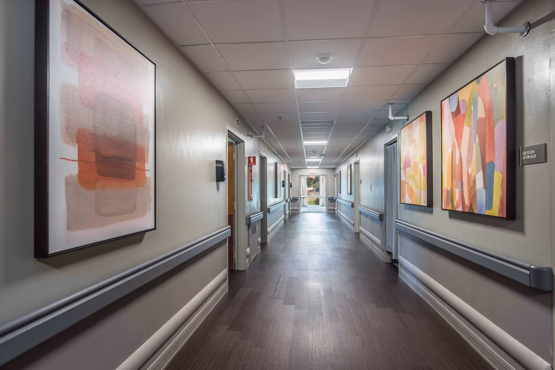 A long, well-lit hallway in a senior living facility with handrails on both sides and abstract artwork hanging on the walls. The floor is dark wood, and there is an exit door with windows at the far end of the corridor.