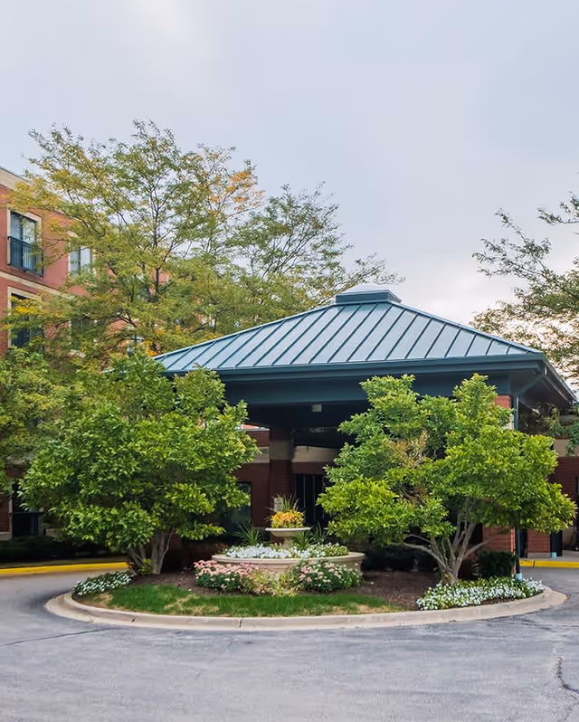Entrance area of Brookdale Vernon Hills facility with a covered driveway, surrounded by green trees and landscaped flower beds under a cloudy sky.
