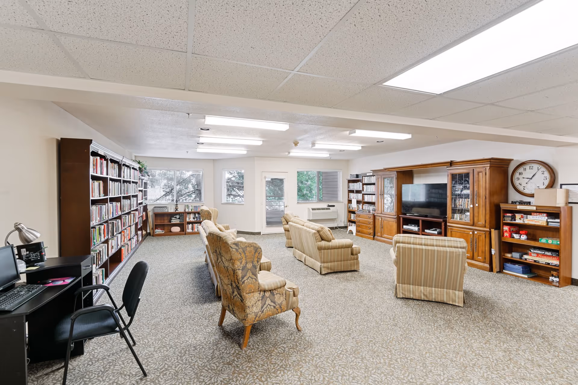 A spacious living room area in Holiday Uffelman Estates featuring multiple upholstered armchairs and sofas arranged facing a large wooden entertainment center with a flat-screen TV. The room has carpeted flooring, fluorescent ceiling lights, and several large windows letting in natural light. There are bookshelves filled with books along one wall and a small desk with a computer and chair in the corner.