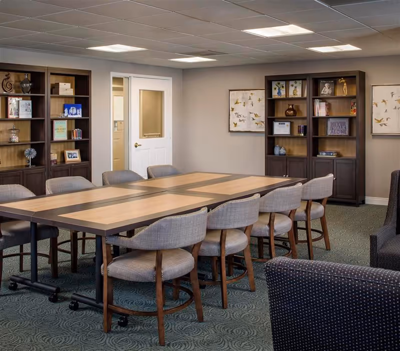 A meeting or activity room with a large rectangular wooden table surrounded by cushioned chairs on wheels. The room has carpeted flooring, beige walls, and a white door with a window. Two dark wood bookshelves with decorative items and framed pictures are against the walls, along with two framed artworks featuring birds.