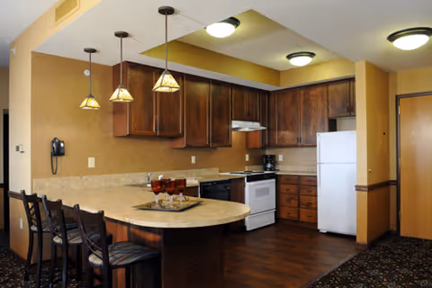 Interior view of a kitchen area with wooden cabinets, a white refrigerator, stove, and dishwasher. There is a curved countertop with four high chairs and three pendant lights hanging above it. The floor is wooden and the walls are painted beige.