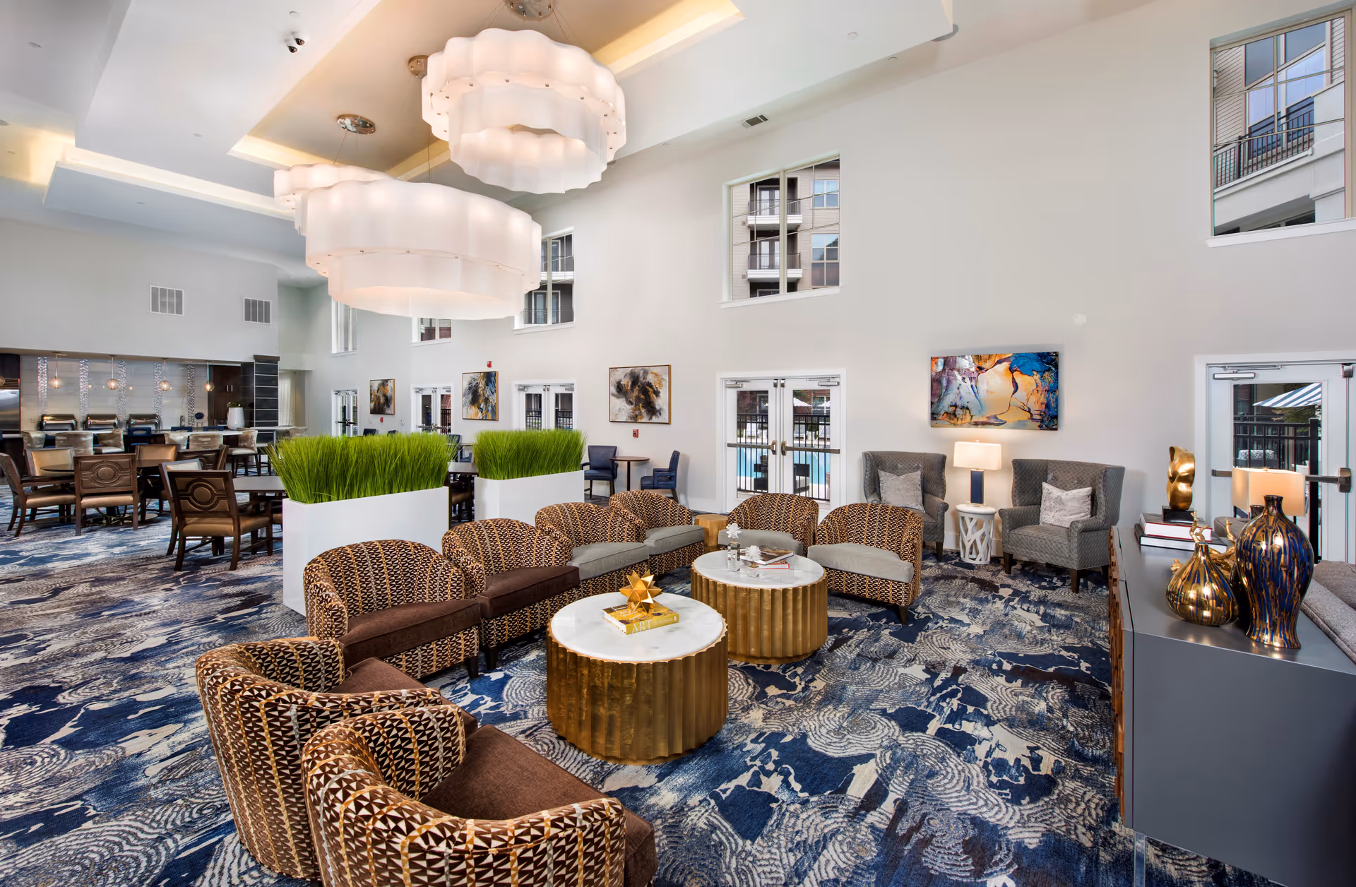 A spacious and modern common area in a senior living facility featuring a semi-circle arrangement of patterned armchairs around two round gold and white marble coffee tables. The room has high ceilings with large, decorative hanging lights, blue and white patterned carpet, and large windows showing an outdoor pool area. There are additional seating areas with chairs and tables, decorative plants, abstract wall art, and a sideboard with vases and sculptures.