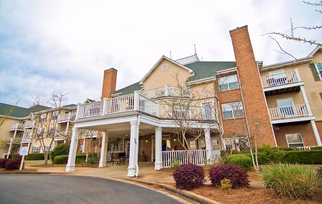 Exterior view of a multi-story assisted living facility with a covered entrance supported by white columns, brick chimneys, balconies, and landscaped bushes and trees in front.