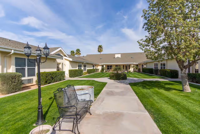 Outdoor courtyard area of a senior living facility with a paved walkway, green grass, bushes, a tree, a black metal bench, and a vintage-style street lamp. The building surrounds the courtyard with beige walls and multiple windows under a clear blue sky.
