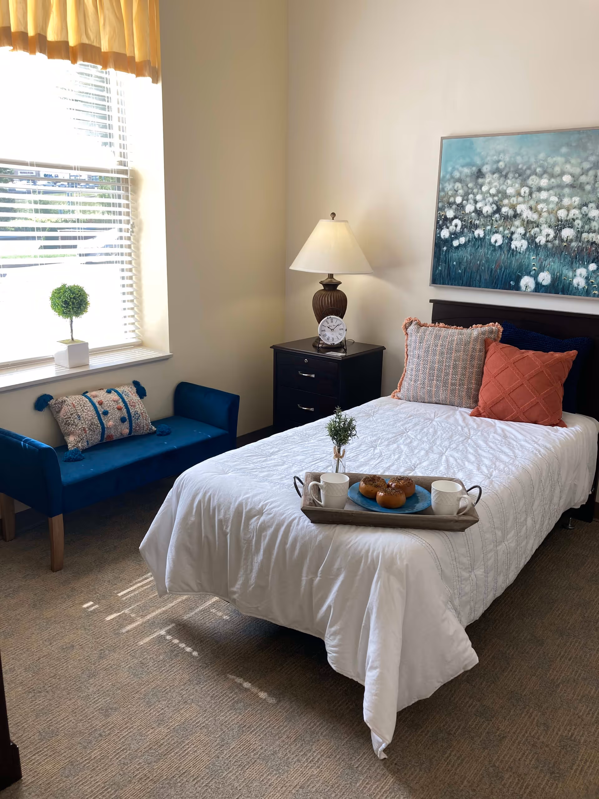 A cozy bedroom with a single bed covered in a white quilt and decorated with three pillows in shades of orange and blue. A wooden nightstand with a lamp and a clock sits beside the bed. A small blue upholstered bench with a decorative pillow is placed under a window with blinds and a yellow valance. A tray with two mugs, a small plant, and three donuts is on the bed. A painting of white flowers on a blue background hangs above the bed.