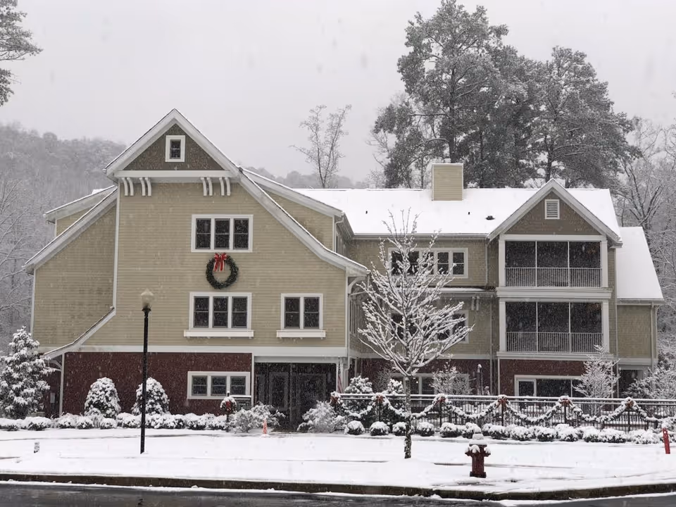 Exterior view of a multi-story residential building covered in snow with a wreath decorated with a red bow hanging on the front. Snow-covered trees and shrubs surround the building, and a snow-covered street and sidewalk are visible in the foreground.