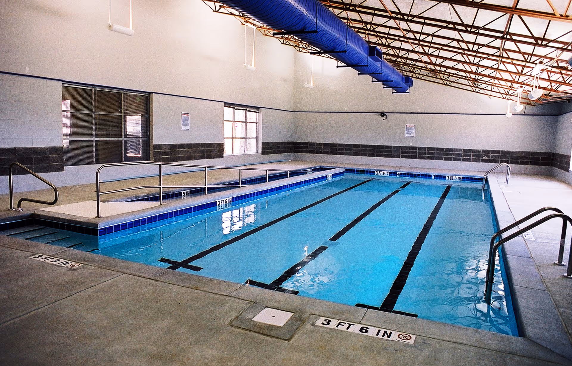 Indoor lap swimming pool with lane markings, handrails, and depth markings inside a bright pool room.