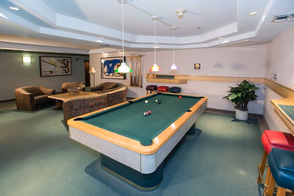 Interior view of a recreational room featuring a pool table with balls arranged on it, three colorful hanging lights above the table, a seating area with patterned sofas and a coffee table, a plant in the corner, and a rack with pool cues on the wall.