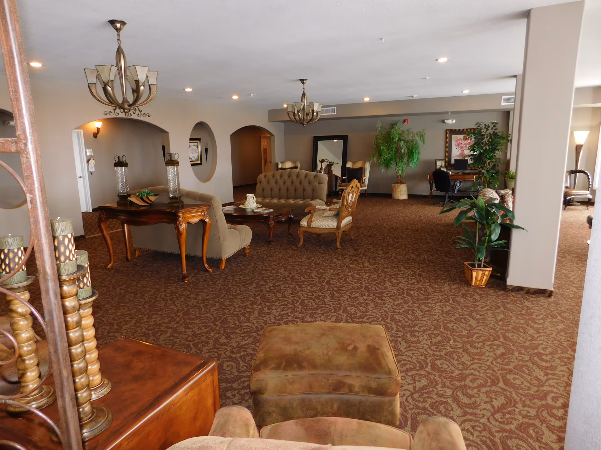 A spacious living room area with patterned carpet, multiple upholstered chairs and sofas, wooden tables, decorative plants, and two chandeliers hanging from the ceiling. There is a desk with a computer and chair in the background, along with framed artwork on the walls.