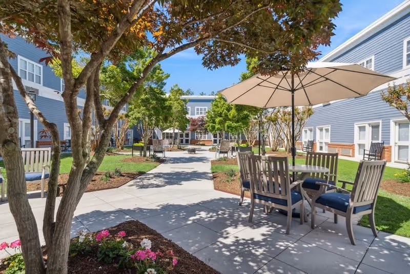Outdoor courtyard area at a senior living facility with paved walkways, trees, flower beds, and seating areas including tables with umbrellas and chairs. The buildings surrounding the courtyard are two-story with blue siding and white trim.