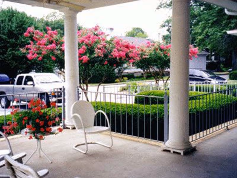 A covered outdoor patio area with white metal chairs and a small table holding a pot of red flowers. The patio overlooks a garden with neatly trimmed hedges and blooming pink flowering trees. There are parked vehicles and houses visible in the background.