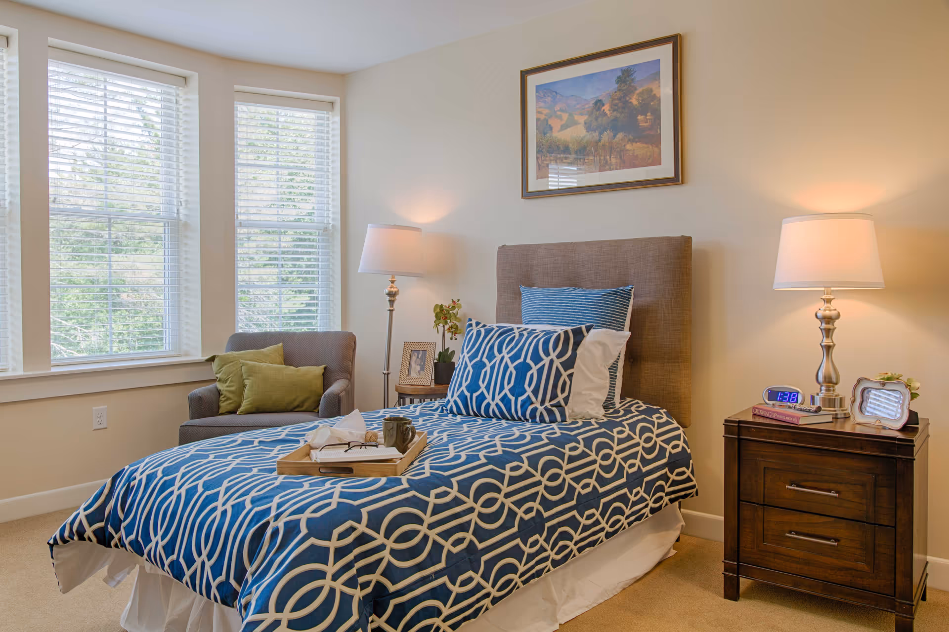Bright bedroom featuring a single bed with blue patterned bedding, a bedside table with a lamp and alarm clock, and an armchair by large windows.