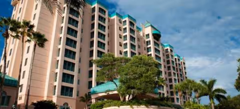 Tall beige senior living building with teal roof accents, palm trees, and blue sky