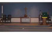 Small chapel interior with an altar, wooden cross on a blue wall, lectern, candles, and seating.
