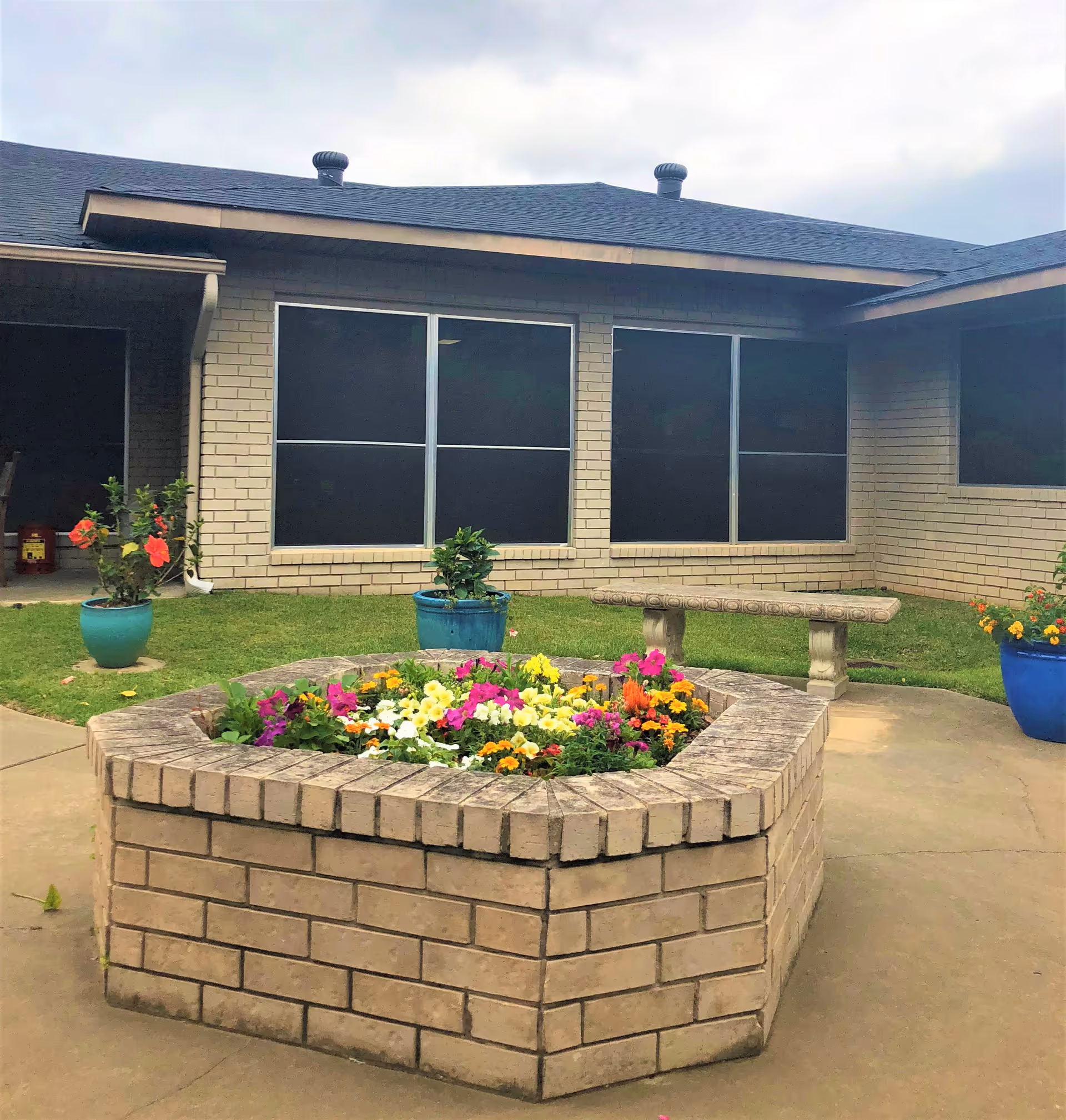 Outdoor courtyard area with a raised brick flower bed filled with colorful flowers, a stone bench, potted plants, and a building with large windows in the background under a cloudy sky.