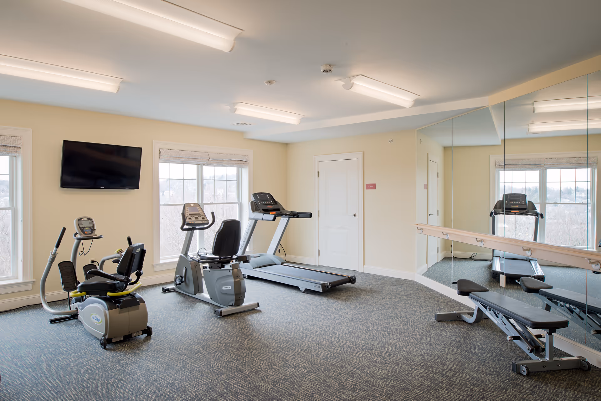 A fitness room with exercise equipment including a recumbent bike, an upright bike, a treadmill, and a weight bench. The room has large windows, a wall-mounted TV, mirrored walls, and overhead fluorescent lighting.