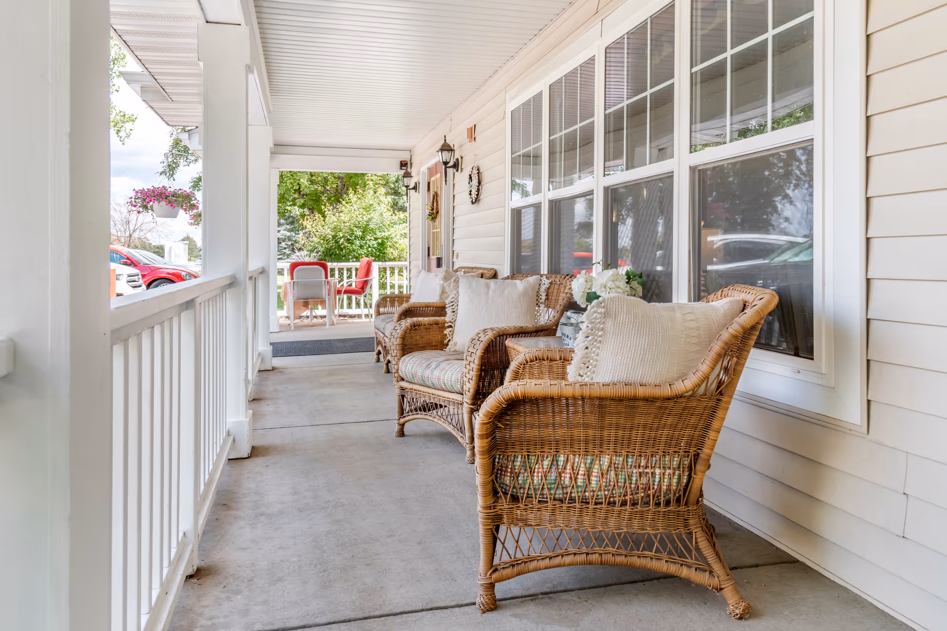 A covered porch area with wicker chairs featuring cushions and pillows, a small table with flowers, white railing, and large windows along the side of the building. In the background, there is a table with red chairs and greenery surrounding the porch.