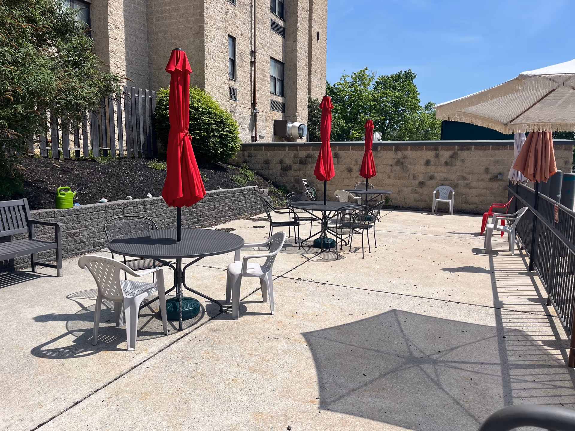 Outdoor patio area with several round metal tables, each with a closed red umbrella in the center. White and black plastic chairs are arranged around the tables. There is a black bench on the left side and a large white umbrella casting a shadow on the concrete ground. The patio is adjacent to a beige brick building with greenery and a wooden fence in the background under a clear blue sky.