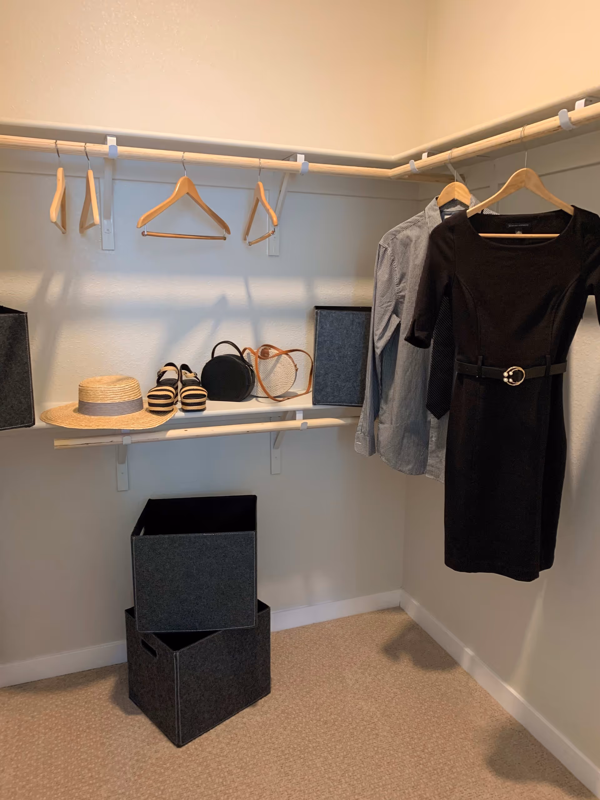 A walk-in closet with beige walls and carpeted floor. The closet has a hanging rod with wooden hangers, some holding a black dress, a blue shirt, and a tie. On a shelf below the rod, there is a straw hat, a pair of striped shoes, and two handbags. Two gray storage bins are stacked on the floor beneath the shelf.