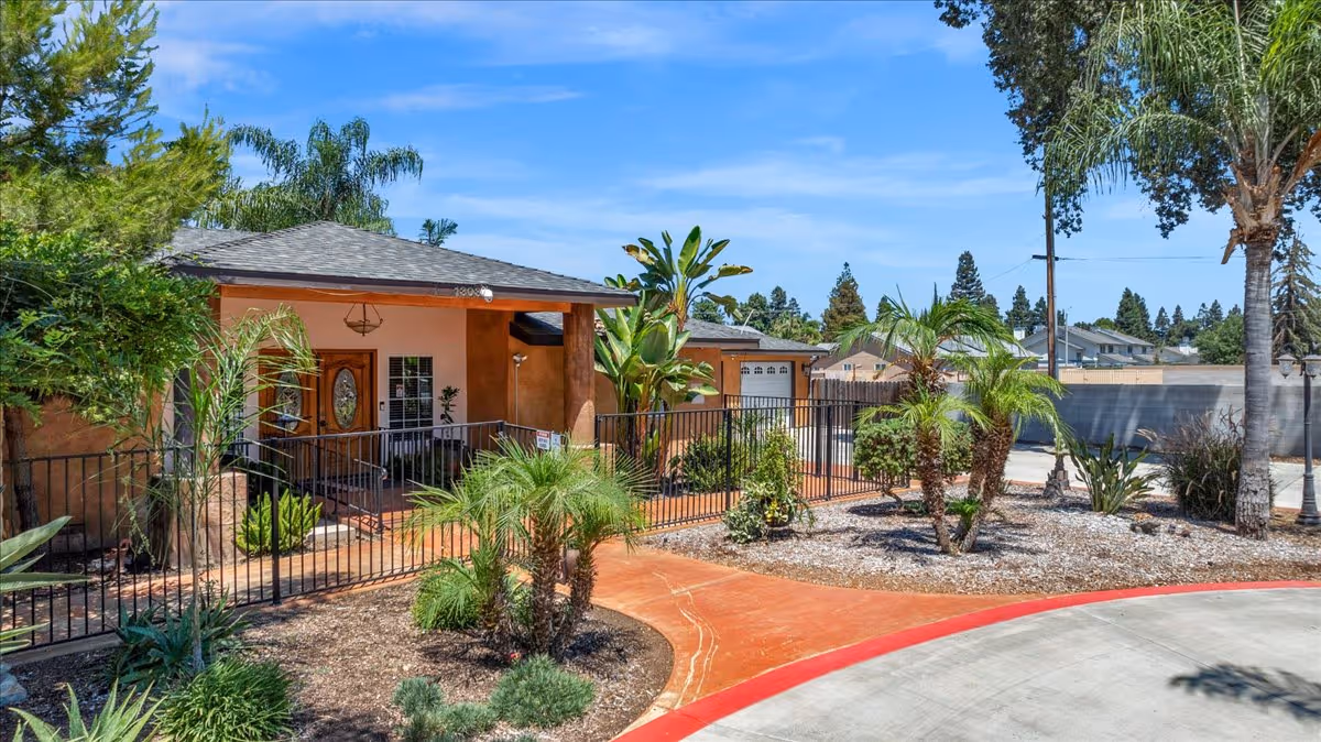 Exterior view of a single-story assisted living facility with a covered entrance, surrounded by a black metal fence and landscaped with palm trees and other greenery under a blue sky.