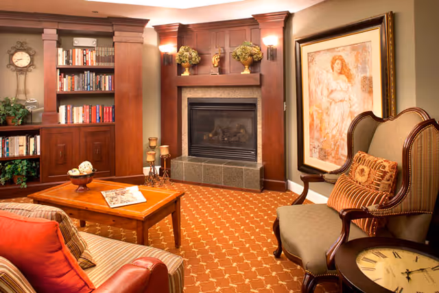 A cozy living room with a fireplace surrounded by wooden paneling and built-in bookshelves filled with books. There is a wooden coffee table with decorative items and a newspaper on it, a striped sofa with red and brown cushions, and an upholstered armchair with a patterned pillow. A large framed painting hangs on the wall next to the armchair, and a round side table with a clock face design is beside the chair. The room has warm lighting and a patterned carpet.