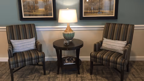 Two striped armchairs with small rectangular pillows are placed on either side of a round wooden side table with a decorative lamp on top. Behind the chairs, there are two framed landscape paintings hanging on a light blue and beige wall with white trim.