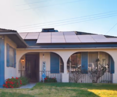 Front exterior view of a single-story house with a tiled roof featuring solar panels. The house has arched windows with blue shutters and a small garden with red flowers in front. A vertical sign near the entrance reads 'WELCOME'.