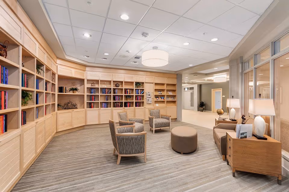 A bright and spacious lounge area with light wood built-in bookshelves filled with books and decorative items. The room features four upholstered armchairs arranged around a round ottoman on a striped carpet. There are two wooden side tables with lamps on the right side near large windows, and the ceiling has recessed lighting and a central round pendant light.